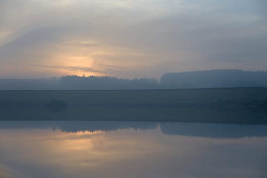 Gentle orange sunset silhouettes a coniferous forest. Dam wall divides the image in half, and the lower half of calm water reflects the sky above.