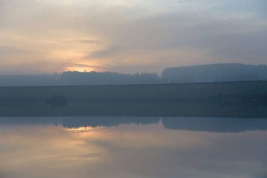 Golden sunset masked by haze and clouds is a back drop to 12 distant figures walking along redmires reservoirs dam wall. Cold day at the end of fall.