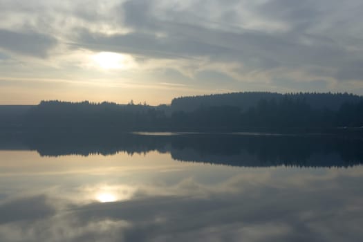 Winters sun and a cloudy sky reflected perfectly in the calm water of redmires reservoir. Hazy forest tree line in the distance, cars parked beneath