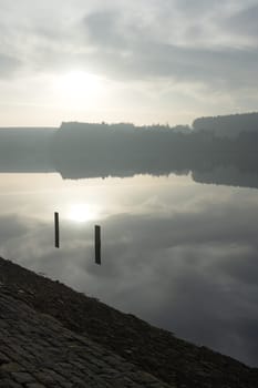 Two posts frame the winters sun as it is reflected in the calm water of a south yorkshire reservoir. Forest background and dam wall foreground.
