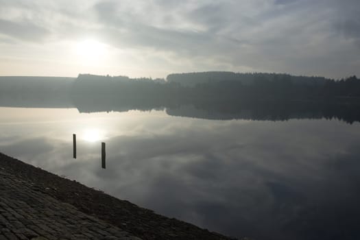 Large reservoir reflects the sky at sunset during winter. A distant forest of conifers is also mirrored in the shimmering water. Tranquil setting