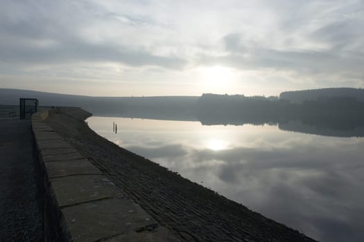 The top dam wall of redmires reservoirs in the peak district. Gently curving around calm water at sunset. Winter time sun and cloudscape is reflected