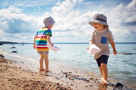 Girl and boy playing on the beach in summer hats and holding paper ships. Happy children running at seaside