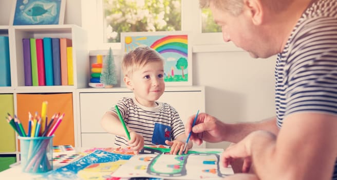 Little child drawing on the paper with father. Boy sitting on the chair at table with paints with dad