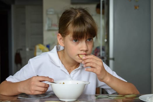 Tired hungry schoolgirl eating soup at the table in the kitchen