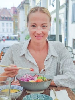 Woman eating tasty colorful healthy natural organic vegetarian Hawaiian poke bowl using asian chopsticks on rustic wooden table. Healthy natural organic eating concept.