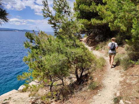 Young active feamle tourist wearing small backpack walking on coastal path among pine trees looking for remote cove to swim alone in peace on seaside in Croatia. Travel and adventure concept.