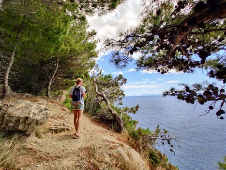 Young active feamle tourist wearing small backpack walking on coastal path among pine trees looking for remote cove to swim alone in peace on seaside in Croatia. Travel and adventure concept.