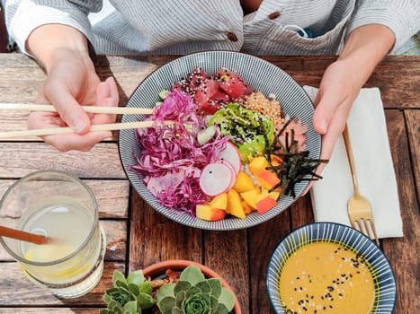 Woman eating tasty colorful healthy natural organic vegetarian Hawaiian poke bowl using asian chopsticks on rustic wooden table. Healthy natural organic eating concept.