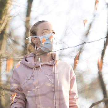Portrait of caucasian sporty woman wearing medical protection face mask while walking in park, relaxing and listening to music. Corona virus, or Covid-19, is spreading all over the world.