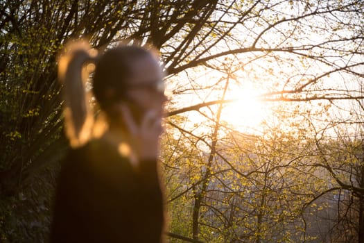 Backlit defocused rear view of young woman talking on cell phone outdoors in park at sunset. Girl holding mobile phone, using digital device, looking at setting sun.