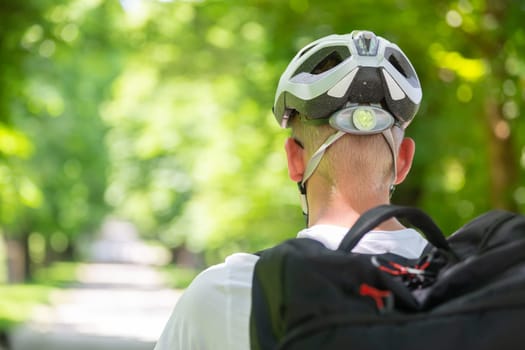 Rear view of unrecognizable man wearing protective helmet on bicycle cycling in city park.