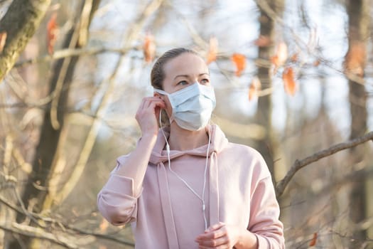 Portrait of caucasian sporty woman wearing medical protection face mask while walking in park, relaxing and listening to music. Corona virus, or Covid-19, is spreading all over the world.
