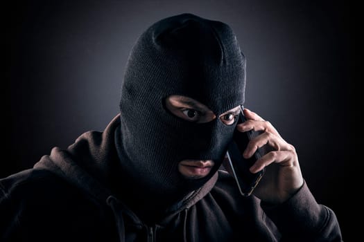Criminal wearing black balaclava and hoodie with a smartphone in the dark