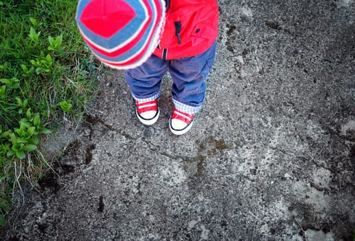 Funny cute boy looking onto own feet. Baby standing on saphalt in red shoes