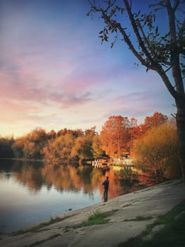 Idyllic sunset scene with a fisher man silhouette using a fishing rod and relaxing at the lake in the park. Beautiful autumn season with colorful trees reflecting on the calm water surface.
