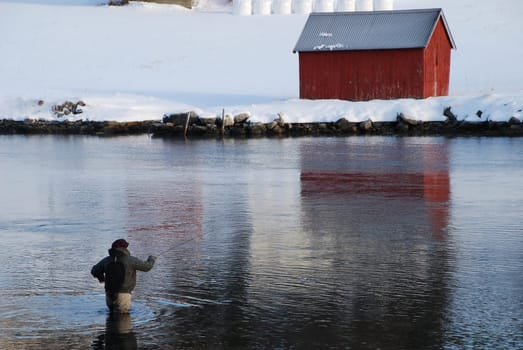 A fly fisherman in waders in Norwegian waters. In front of him the snow-covered area and a typical Norwegain red wodden house.