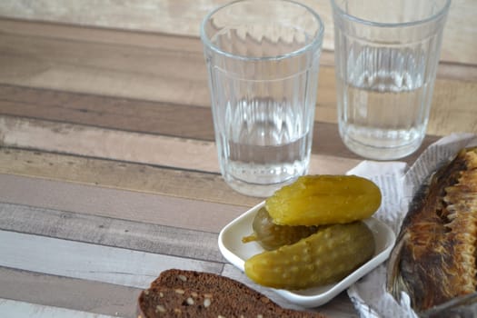 Shot of vodka with a stockfish and pickled cucumber on an old wooden table. Copy space