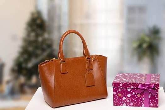 Template for christmas sale. Elegant brown female luxury handbag and a gift box on table against blurred xmas tree background indoor. Advertising fashionable woman accessories.