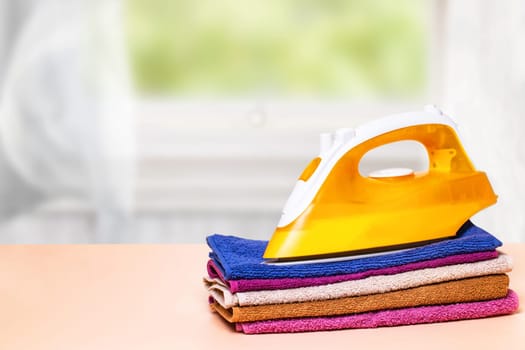 Closeup of a yellow iron on a stack colorful towels on table against an abstract bright curtain backgound. Advertising for the laundry service with space for design.