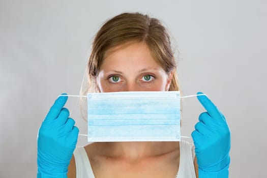 Woman stretching ear loops of blue surgical mask as she is attaching it to her face. Nurse protecting her respiratory system against coronavirus. Horizontal portrait of a female.