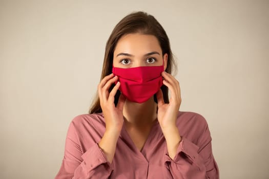Young woman touching red mask on the face in studio. Girl with brown hair wearing textile respiratory like preventions against virus. Brunette female with covered mouth looking to the camera.