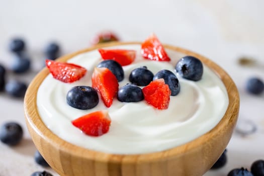 Wooden bowl filled with yoghurt, strawberries and blueberries in detail. Little fresh berries in white cream on table. Woody dish full of dairy product with red and blue fruits in focus.