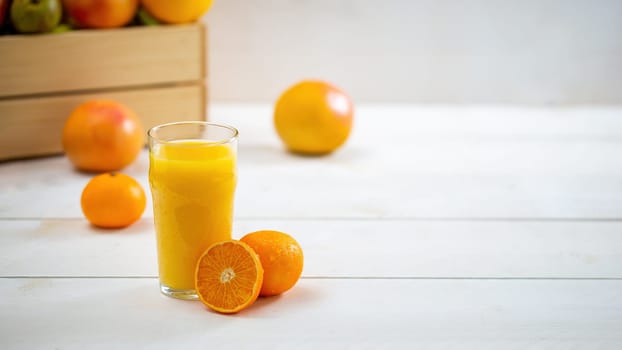Glass of juice with cut orange on white desk with copy space. Cup of fresh yellow bevarage with grapefruits and wooden box in the background. Still life of sweet healthy drink with fruit with space for text.