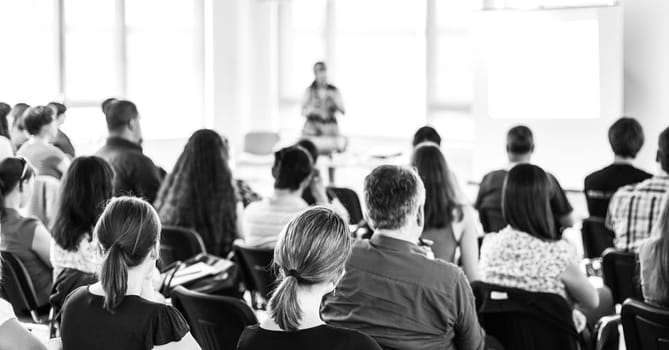 Business and entrepreneurship symposium. Female speaker giving a talk at business meeting. Audience in conference hall. Rear view of unrecognized participant in audience.