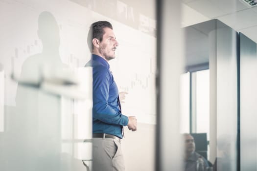 Business man making a presentation in front of whiteboard. Business executive delivering a presentation to his colleagues during meeting or in-house business training. View through glass.