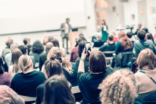 Male speaker giving presentation in lecture hall at university workshop. Audience in conference hall. Rear view of unrecognized participant in audience. Scientific conference event.