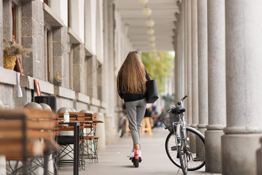 Rear view of girl riding public rental electric scooter in urban city environment at fall. New eco-friendly modern public city transport in Ljubljana.