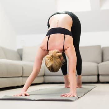 Beautiful blonde woman doing home workout indoors. Woman practice yoga at home. Fit girl using workout tutorials for healthy active lifestyle. Woman using quarantine for home workouts. Stretching