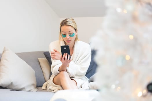 Woman wearing cosy warm bathrobe and cosmetic eye patches relaxing on sofa using social media on phone for video chatting with her loved ones in winter Christmas time.