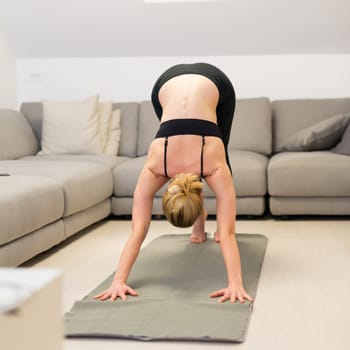 Beautiful blonde woman doing home workout indoors. Woman practice yoga at home. Fit girl using workout tutorials for healthy active lifestyle. Woman using quarantine for home workouts. Stretching