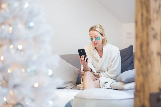 Woman wearing cosy warm bathrobe and cosmetic eye patches relaxing on sofa using social media on phone for video chatting with her loved ones in winter Christmas time.