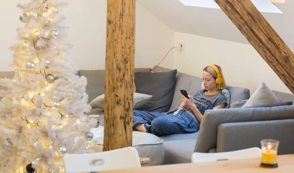 Young thoughtful woman sitting indoors at home living room sofa using social media on phone for video chatting and staying connected with her loved ones. Stay at home, social distancing lifestyle.