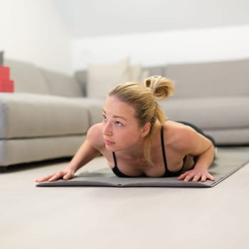 Beautiful blonde woman doing home workout indoors. Woman practice yoga at home. Fit girl using workout tutorials for healthy active lifestyle. Woman using quarantine for home workouts. Stretching