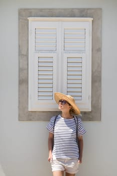 Beautiful young female tourist woman wearing sun hat, standing and relaxing in front of vinatage wooden window in old Mediterranean town while sightseeing on hot summer day.