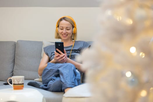 Young cheerful woman sitting indoors at home living room sofa using social media on phone for video chatting and staying connected with her loved ones. Stay at home, social distancing lifestyle.