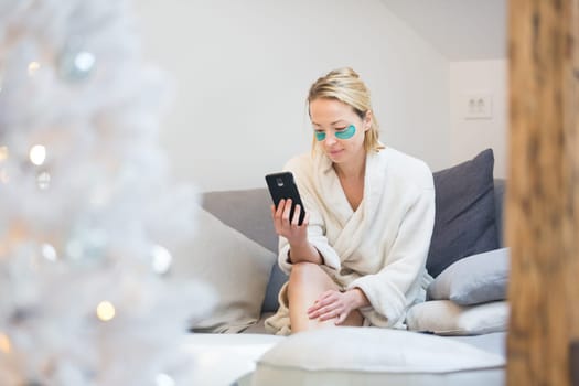 Woman wearing cosy warm bathrobe and cosmetic eye patches relaxing on sofa using social media on phone for video chatting with her loved ones in winter Christmas time.