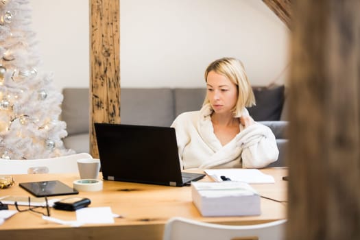 Young businesswoman wearing cosy warm bathrobe working remotly from home in winter Christmas time during 2020 corona virus pandemic. Work from home, selter in place, concept.