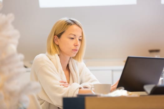 Young businesswoman wearing cosy warm bathrobe working remotly from home in winter Christmas time during 2020 corona virus pandemic. Work from home, selter in place, concept.