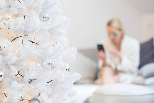 Defocused image of young cheerful woman sitting indoors at christmas decorated home living room sofa using social media on phone for video chatting and staying connected with her loved ones.