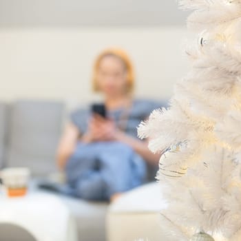 Defocused image of young cheerful woman sitting indoors at home living room sofa using social media on phone for video chatting and staying connected with her loved ones.