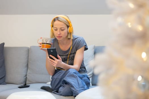 Young cheerful woman sitting indoors at home living room sofa using social media on phone for video chatting and staying connected with her loved ones. Stay at home, social distancing lifestyle.