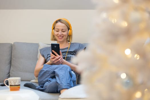 Young cheerful woman sitting indoors at home living room sofa using social media on phone for video chatting and staying connected with her loved ones. Stay at home, social distancing lifestyle.