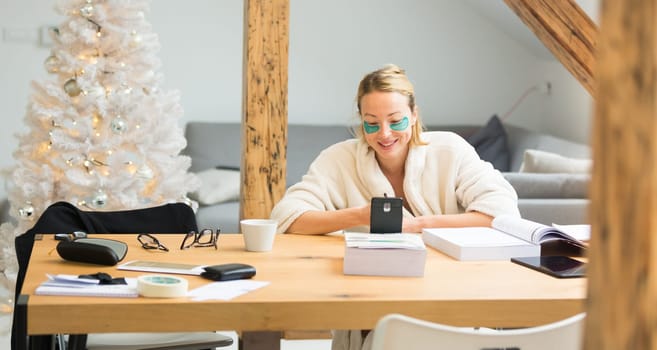 Young businesswoman wearing cosy warm bathrobe and cosmetic eye patches working remotly from home in winter Christmas time during 2020 corona virus pandemic. Work from home, selter in place, concept.