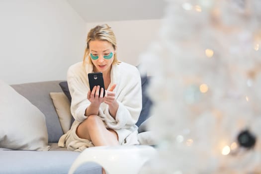 Woman wearing cosy warm bathrobe and cosmetic eye patches relaxing on sofa using social media on phone for video chatting with her loved ones in winter Christmas time.