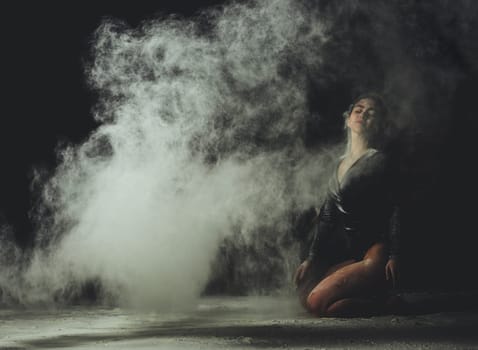 young beautiful caucasian woman with long hair is dressed in a sports black bodysuit and sits on the floor and throwing white flour up, black background
